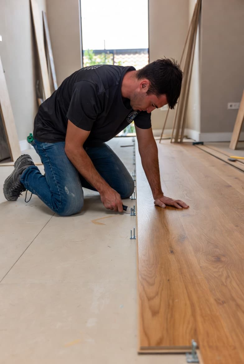 Installer laying wide-plank hardwood flooring with precision spacers along the wall.